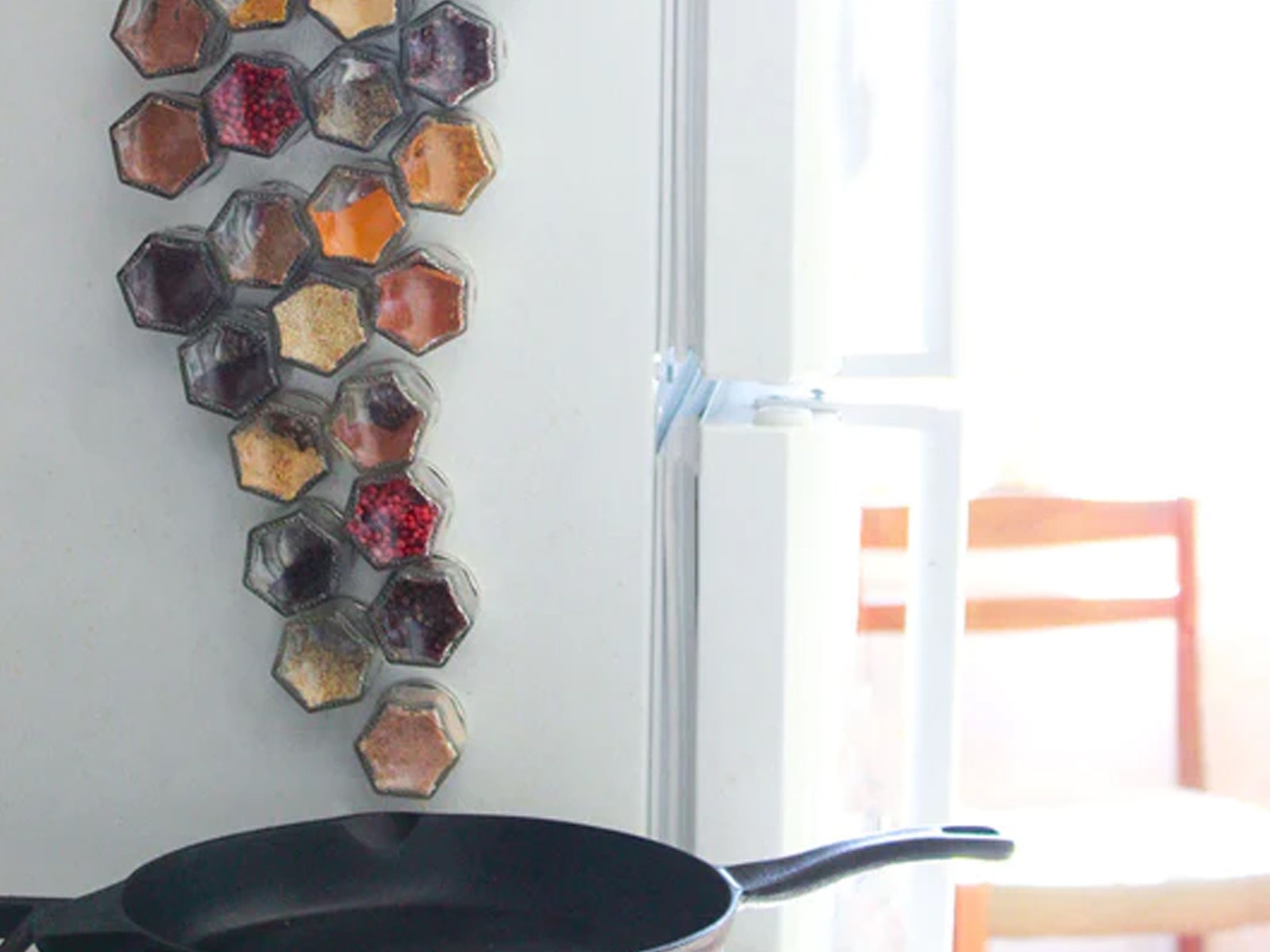 Clear hexagonal spice jars magnetically sticking to the side of a white fridge with a frying pan in immediate view